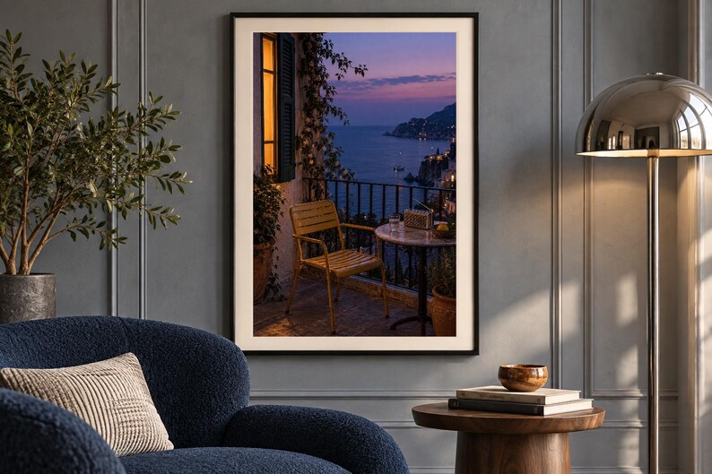 May include: A framed wall art print depicting a balcony scene at dusk, featuring a yellow chair and a small round table overlooking a coastal landscape with a dark blue sea, a purple sunset sky, and a warm glowing window light on the left side.
