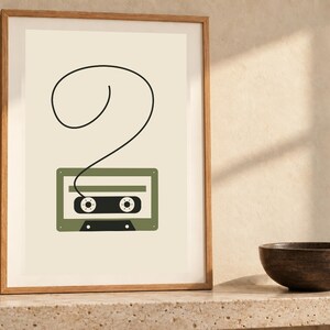 Pode incluir: Uma impress&atilde;o de arte emoldurada apresentando uma ilustra&ccedil;&atilde;o minimalista de uma fita cassete verde-oliva em um fundo creme, com uma &uacute;nica linha preta estendendo-se do topo da fita para formar um grande ponto de interroga&ccedil;&atilde;o em la&ccedil;o.