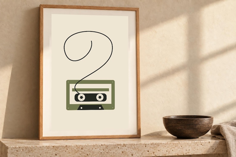Pode incluir: Uma impress&atilde;o de arte emoldurada apresentando uma ilustra&ccedil;&atilde;o minimalista de uma fita cassete verde-oliva em um fundo creme, com uma &uacute;nica linha preta estendendo-se do topo da fita para formar um grande ponto de interroga&ccedil;&atilde;o em la&ccedil;o.