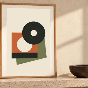 May include: A framed wall art print featuring a geometric composition on a cream background. The design includes a black circular ring, an orange square with a white circle cutout, a solid black rectangle, and an olive green rectangular shape.
