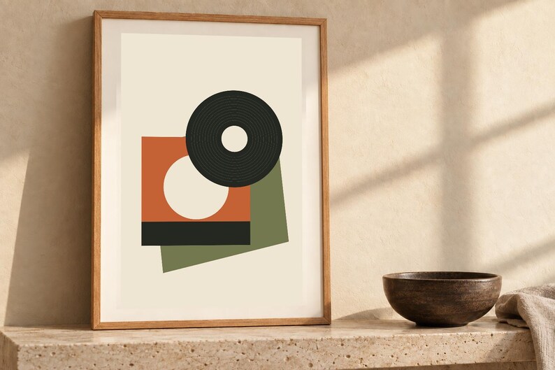 May include: A framed wall art print featuring a geometric composition on a cream background. The design includes a black circular ring, an orange square with a white circle cutout, a solid black rectangle, and an olive green rectangular shape.