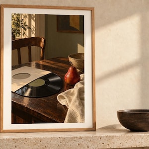 May include: A framed wall art print featuring a photograph of a dark wooden table with a black vinyl record in a sleeve, a red pear, a textured cloth, and a wooden bowl, all illuminated by warm sunlight casting long shadows across the scene.