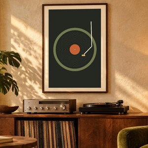 May include: A framed wall art print featuring a graphic illustration of a vinyl record player. The design displays a dark gray background, a muted green circular record, a central orange label, and a white tone arm line.