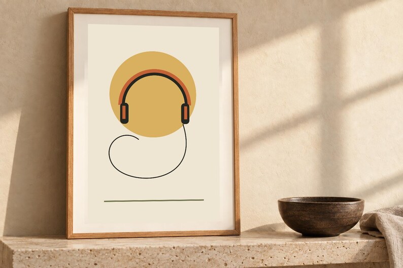 May include: A framed wall art print featuring a minimalist graphic of black and orange headphones centered over a large yellow circle on a cream background, with a thin black line forming a loop below and a straight horizontal line at the base.
