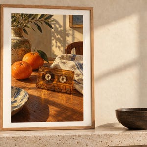 May include: A framed wall art print featuring a wooden table with two bright orange citrus fruits, a translucent amber cassette tape, a ceramic vase with green leaves, and a white cloth with blue stripes, all captured in warm sunlight.
