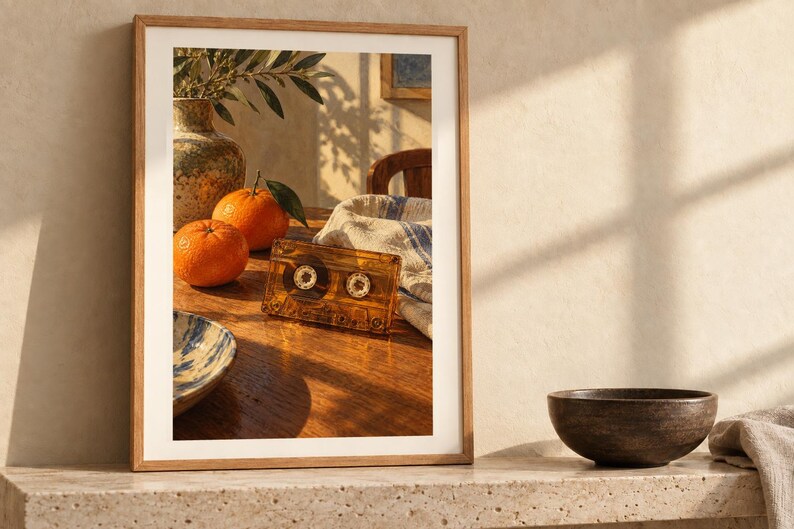 May include: A framed wall art print featuring a wooden table with two bright orange citrus fruits, a translucent amber cassette tape, a ceramic vase with green leaves, and a white cloth with blue stripes, all captured in warm sunlight.