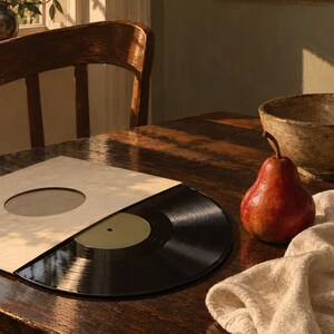 May include: A black vinyl record partially pulled from a light-colored paper sleeve with a circular cutout. A single red pear sits next to the record on a dark wooden surface, accompanied by a textured off-white fabric cloth nearby.