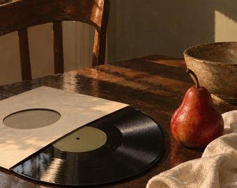 May include: A black vinyl record partially pulled from a light-colored paper sleeve with a circular cutout. A single red pear sits next to the record on a dark wooden surface, accompanied by a textured off-white fabric cloth nearby.