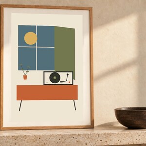 May include: A framed wall art print featuring a graphic illustration of a record player on an orange console table. Above the table is a blue window with a yellow sun and a small potted plant. The print is set against a cream background.