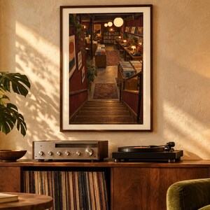 May include: A framed wall print depicting a staircase leading down into a dimly lit record store with rows of vinyl albums, warm pendant lighting, and patterned rugs on the floor, displayed above a wooden media console with audio equipment.