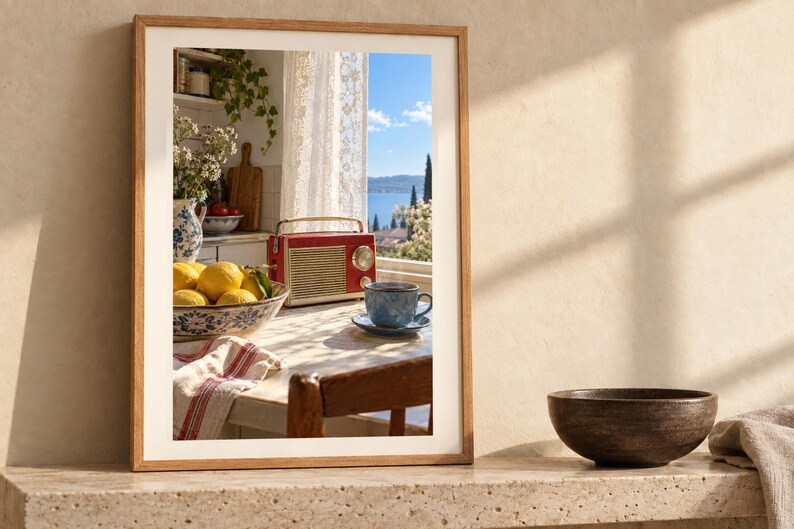 May include: A framed wall art print featuring a kitchen scene with a red radio, a bowl of yellow lemons, and a blue teacup on a table. The view through the window shows a blue sea and cypress trees. The frame is light wood with a white mat border.
