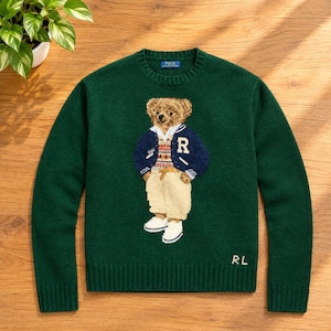 Könnte beinhalten: Ein dunkelgrüner Langarmpullover mit Rundhalsausschnitt und einem Teddybären-Motiv, der eine marineblaue College-Jacke mit dem Buchstaben R, eine gemusterte Weste und eine beige Hose trägt. Die Buchstaben RL sind unten rechts in Weiß eingestickt.