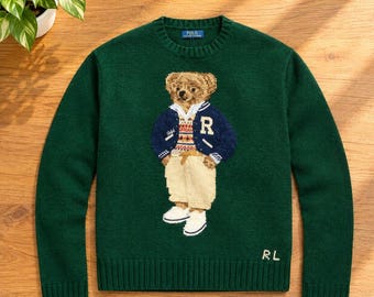Può includere: Un maglione a maniche lunghe girocollo verde scuro con un'immagine centrale di un orsacchiotto che indossa una giacca varsity blu navy con la lettera R, un gilet a motivi geometrici e pantaloni beige. Le lettere RL sono ricamate in bianco in basso a destra.