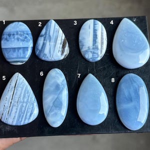 May include: Eight polished blue opal cabochons arranged on a black display board, numbered one through eight. The stones feature various shapes including oval and teardrop, displaying light blue tones with white banding and dark inclusions.