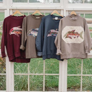 May include: Four long-sleeve sweatshirts in burgundy, olive, navy, and taupe. The first three feature a floral fabric fish appliqué. The taupe sweatshirt has a textured, cream-coloured oval patch with a floral fabric fish design stitched onto the front.