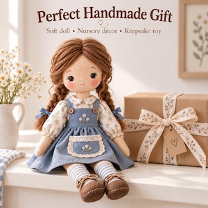 May include: A soft doll with brown yarn hair in two braids, wearing a floral print blouse under a blue denim-style pinafore dress with a white lace-trimmed pocket. The doll has striped socks and brown shoes, positioned next to a gift box.