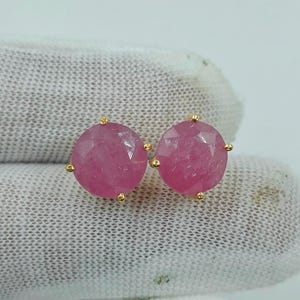 Puede incluir: Un par de pendientes de botón con piedras preciosas rosas redondas y facetadas, montadas en garras de metal dorado. Cada pendiente presenta una piedra rosa circular y translúcida con una superficie multifacetada que refleja la luz.