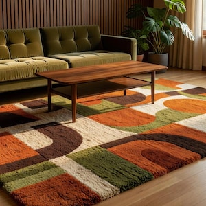 Può includere: Un tappeto rettangolare caratterizzato da un motivo geometrico di archi e blocchi intrecciati nei toni dell'arancione bruciato, verde oliva, marrone scuro e crema. Il tappeto ha un pelo spesso e strutturato che copre il pavimento in legno.