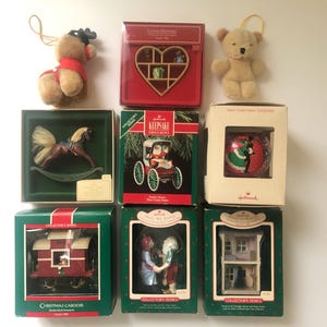 May include: A collection of nine Hallmark Keepsake Christmas ornaments. The ornaments are in boxes and feature a variety of themes, including a rocking horse, a carousel, a teddy bear, a heart, and a Christmas village. The boxes are all red and white with the Hallmark logo.