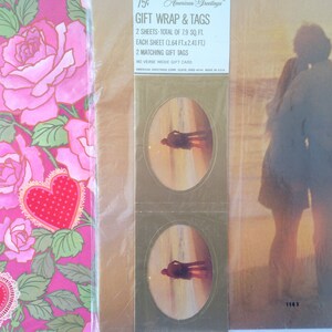 May include: Gift wrap and tags set. The gift wrap features a pink rose and heart design. The tags have an oval image of a couple on a beach. The packaging includes the text "Gift Wrap & Tags" and "American Greetings".