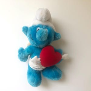 May include: A blue plush Smurf holding a red heart with an arrow through it. The Smurf is wearing a white hat and a white nappy.
