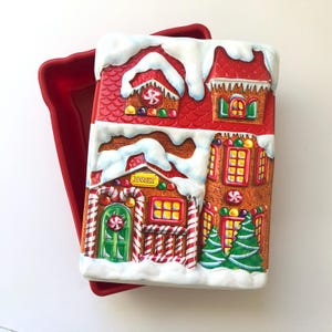 May include: A rectangular gingerbread house-shaped baking mould with a red exterior. The house features a red roof with white snow, colourful candies, and the word "Bakery" above the door. The mould sits in a red baking pan.