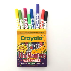 May include: A box of Crayola Mini Stampers washable markers. The box is yellow and purple with the Crayola logo and the words "Mini Stampers" and "Washable" printed on it. The markers are various colors, with stamp designs on the tips.