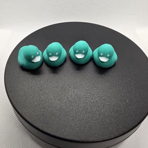 May include: Four small teal rubber ducks arranged in a horizontal row. Each duck features a smooth teal body, a white beak, and two small white eyes. The ducks are identical in size and color, displaying a simple, molded form with rounded features.