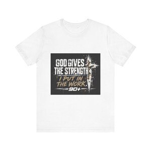 Può includere: Una t-shirt bianca a maniche corte con una grafica rettangolare sul petto. La grafica presenta uno sfondo scuro con il testo bianco "GOD GIVES THE STRENGTH" e "I PUT IN THE WORK" in un carattere usurato, insieme a una croce e "90+".