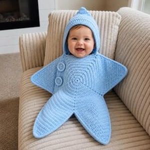 Può includere: Un sacco nanna per neonati all'uncinetto color azzurro a forma di stella a cinque punte. Il capo presenta un cappuccio a punta, un motivo circolare centrale e due grandi bottoni blu abbinati sul lato. La trama è uniforme su tutto il pezzo.