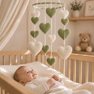 May include: A hanging mobile featuring multiple crocheted hearts in shades of sage green and cream, suspended from a circular frame by white strings. The hearts vary in size and are arranged in a vertical pattern above a wooden crib.