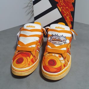 Könnte beinhalten: Ein Paar orange-gelbe Adidas-Sneaker mit einem lebhaften Blumenmuster auf der Zehenkappe, dicken orangefarbenen Schnürsenkeln und einer weißen Zunge mit Adidas-Logo. Ein Etikett am rechten Schuh zeigt AdaMo adisgn.-shoes in einer Sprechblase.