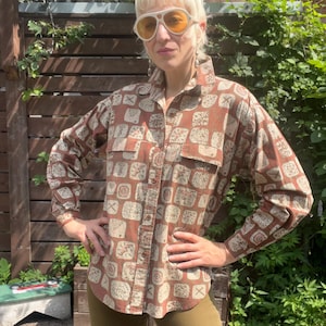 May include: A long-sleeved button-down shirt in a brown and beige geometric square pattern, featuring a pointed collar, two chest pockets, and a curved hemline, paired with solid olive green trousers and large white-framed sunglasses.
