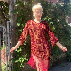 May include: A long-sleeved dress featuring a deep red velvet burnout pattern over a tan base fabric. The garment has a V-neckline and a hemline finished with long, dangling red fringe that extends from the bottom edge of the velvet fabric.