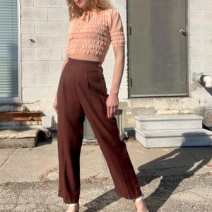 May include: A woman wearing a peach-colored knitted sweater with a high neck and a brown pair of high-waisted pants.