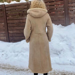 Op de afbeelding: Een lange, beige jas met capuchon en riem, gedragen over een zwarte broek en schoenen. De jas lijkt gemaakt van een zacht materiaal, mogelijk su&egrave;de of schapenvacht. De achtergrond toont een besneeuwd gebied en een houten hek.