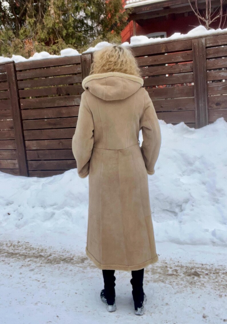 Op de afbeelding: Een lange, beige jas met capuchon en riem, gedragen over een zwarte broek en schoenen. De jas lijkt gemaakt van een zacht materiaal, mogelijk su&egrave;de of schapenvacht. De achtergrond toont een besneeuwd gebied en een houten hek.