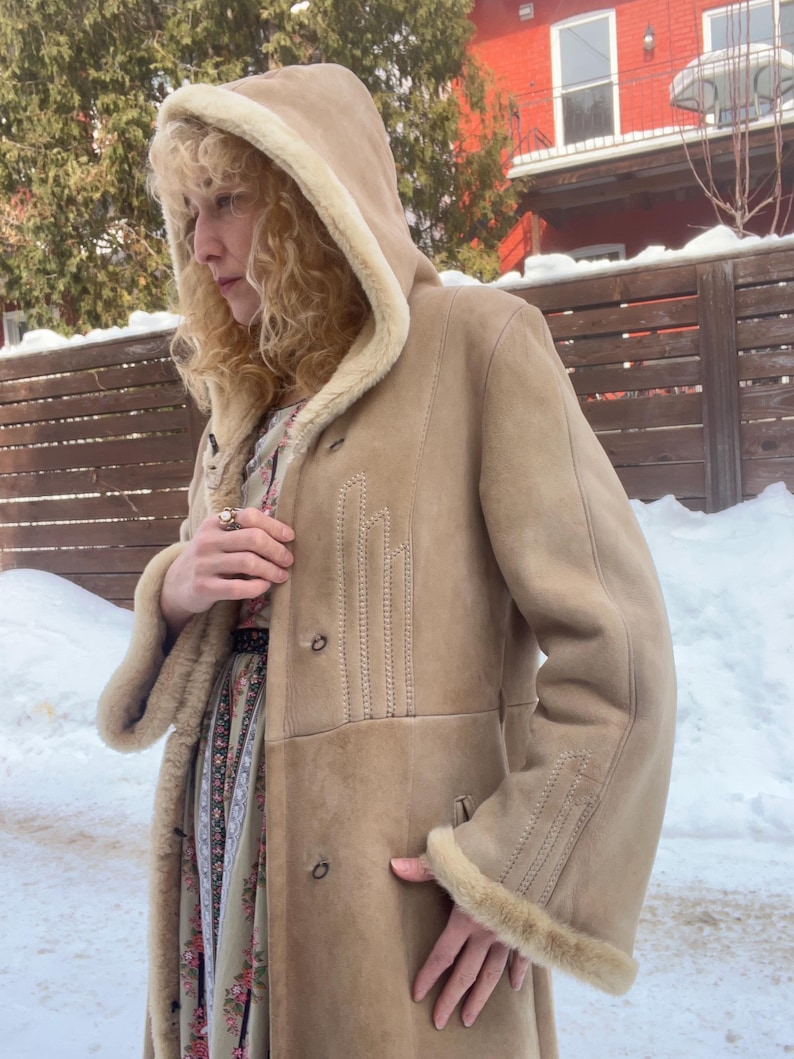 Op de afbeelding: Een bruin su&egrave;de jas met een capuchon gevoerd met cr&egrave;mekleurig imitatiebont. De jas heeft knoopsluitingen en decoratieve stiksels op de voorkant en mouwen. De jas is een lange stijl, geschikt voor koud weer.