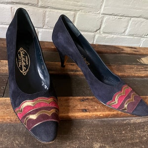 May include: Pair of navy blue suede heels with gold and burgundy trim. The heels have a pointed toe and a stiletto heel. The shoes are labeled "Granada Shoes Collection".