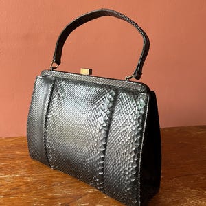 May include: A black handbag with a textured snakeskin pattern, featuring a single arched top handle and a rectangular frame. The bag has a metallic gold-toned clasp closure at the top centre and a structured, tapered silhouette with vertical paneling.