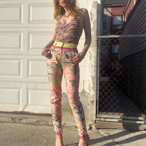 Pode incluir: Uma mulher vestindo uma cal&ccedil;a de estampa floral rosa e branca com um cinto amarelo e um top de estampa de zebra marrom. Ela est&aacute; usando &oacute;culos de sol e saltos dourados.