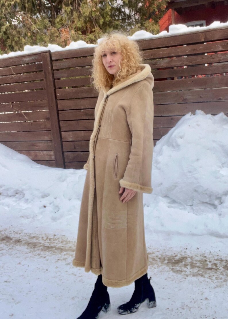 Op de afbeelding: Een lange, bruinbeige jas met een capuchon en een lichtgekleurde voering. De jas heeft een knoopsluiting en wordt gedragen over zwarte laarzen. De persoon staat in een besneeuwde omgeving met een houten hek op de achtergrond.