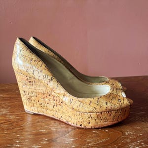 May include: A pair of peep-toe wedge shoes featuring a natural tan cork texture throughout the exterior. The shoes have a high wedge heel, a platform sole, and a light-colored interior lining, showcasing a consistent cork pattern across the entire surface.