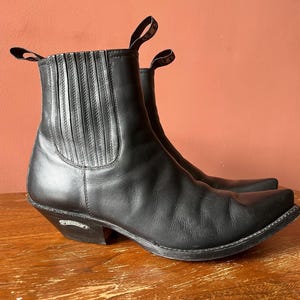 May include: A pair of black leather Chelsea boots featuring a pointed toe, elastic side panels, and a pull tab at the top of the shaft. The boots have a stacked heel with a small silver-toned metal emblem embedded on the outer side of the heel.