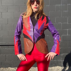 Puede incluir: Una chaqueta de cuero de color morado, marrón y rojo con cierre de cremallera. La chaqueta tiene un patrón geométrico y se lleva sobre una camiseta de cuello alto negra.