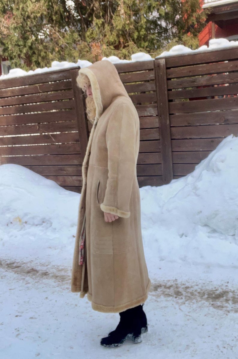 Op de afbeelding: Een lange, bruine jas met een capuchon en bontafwerking. De jas wordt gedragen over zwarte laarzen in een besneeuwde buitenomgeving. De jas heeft een knoopsluiting en zakken.
