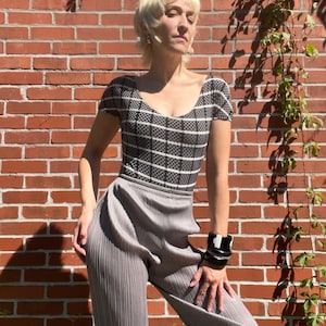 May include: A short-sleeved bodysuit featuring a black and white checkered pattern with a scoop neckline, paired with high-waisted, light gray pleated trousers that have a vertical texture. A black and white stacked bangle bracelet is worn on the wrist.