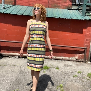 May include: A woman wearing a yellow and green striped dress with red floral accents. The dress has a sleeveless design and a fitted silhouette. She is wearing sunglasses and sandals.