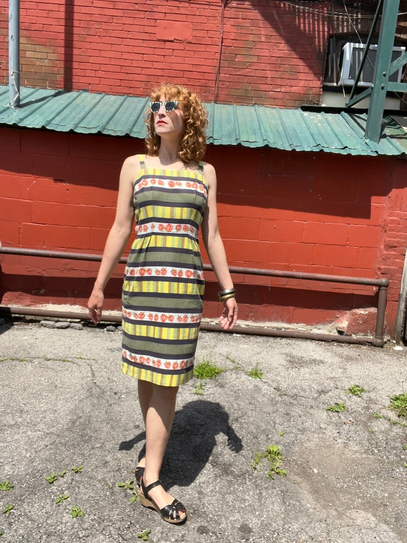 May include: A woman wearing a yellow and green striped dress with red floral accents. The dress has a sleeveless design and a fitted silhouette. She is wearing sunglasses and sandals.
