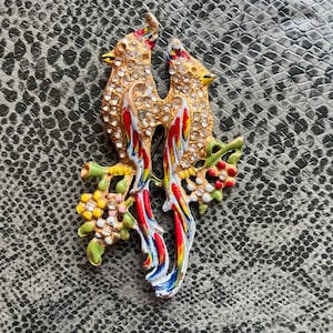 Puede incluir: Broche dorado de pájaros con dos aves enfrentadas, adornado con pedrería transparente. Las aves tienen detalles rojos, azules y amarillos en sus alas y están posadas en ramas verdes con flores coloridas.